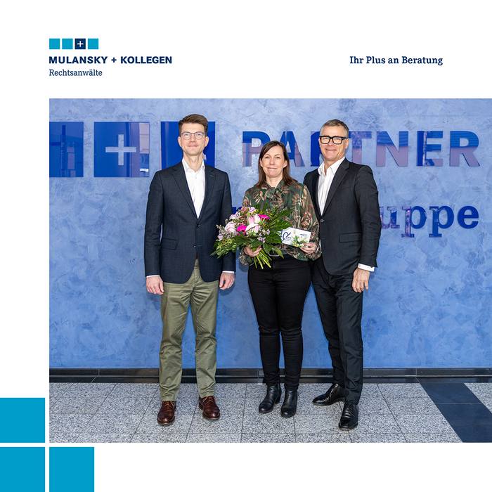 Zwei Männer stehen links und rechts von einer Frau, die einen Blumenstrauß hält.